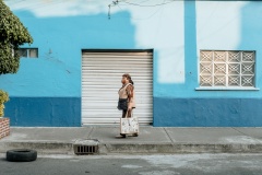 Eine Frau, die Mutter des Fotografen, läuft mit Einkaufstasche und Mundschutz vor einer blauen Wand