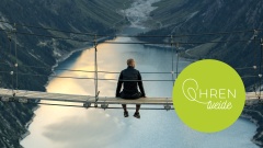 Mann sitzt auf einer Hängebrücke hoch über einem Bergsee
