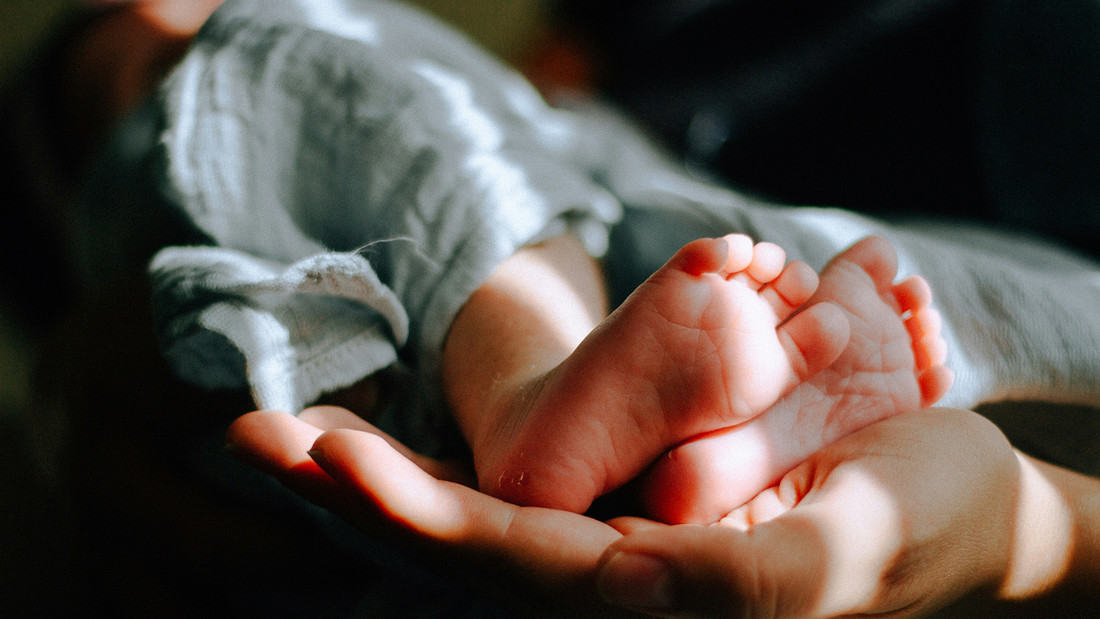 Babyfüße in einem grauen Leinentuch, von einer erwachsenen Hand gehalten
