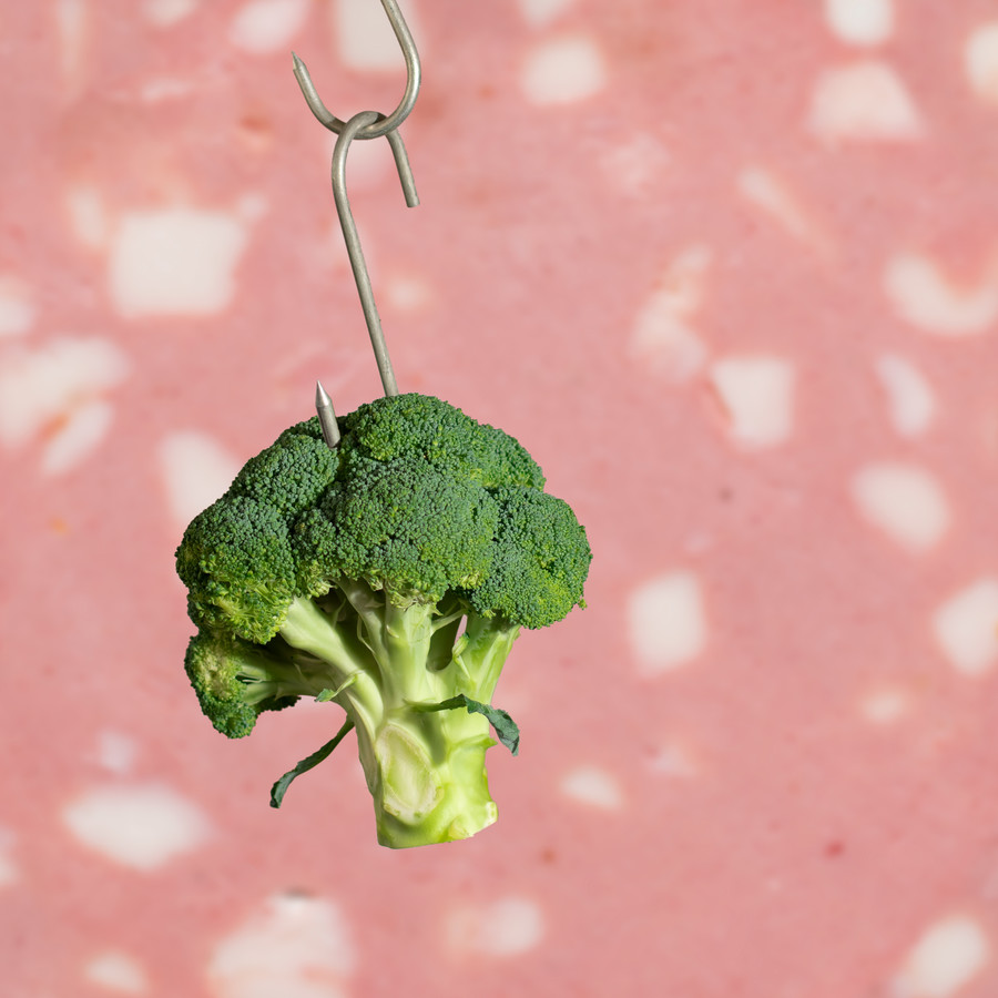 Brokkoli hängt vor einer überdimensionalen Mortadellascheibe an einem Fleischerhaken