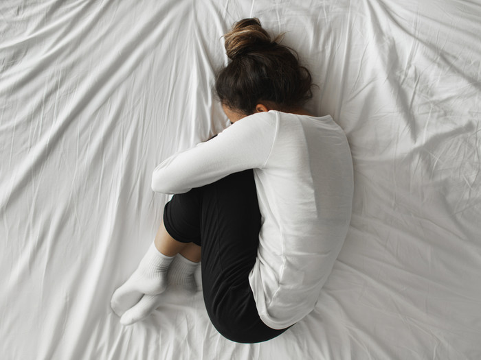 Eine junge Frau liegt in Embryostellung zusammengekrümmt auf einem Bett mit weißen Laken