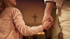 Ein Kind und eine Person Hand in Hand vor einem Kreuz.