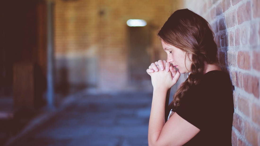 Eine junge Frau lehnt mit dem Rücken an einer Backsteinwand und betet.