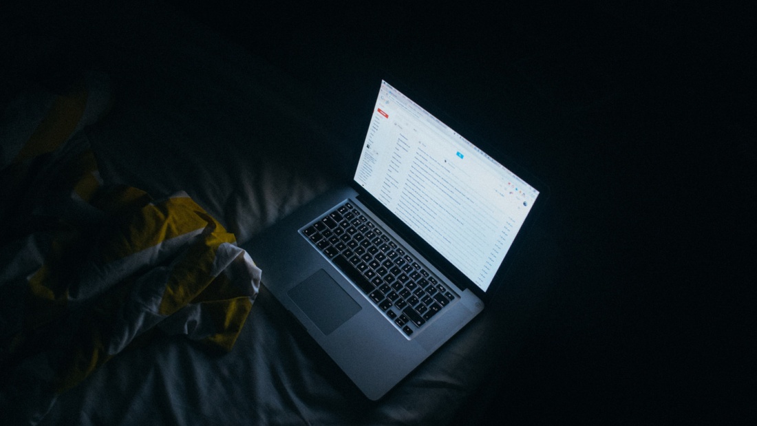 Der Bildschirm eines Laptops leuchtet auf einem Bett in einem dunklen Arbeitszimmer.