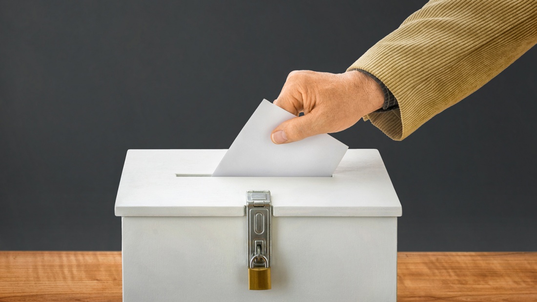 Eine Hand steckt einen Wahlumschlag in eine Box.