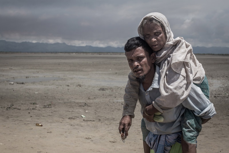 Ein Rohingya trägt eine alte Frau auf dem Rücken. Sie sehen beide erschöpft aus.