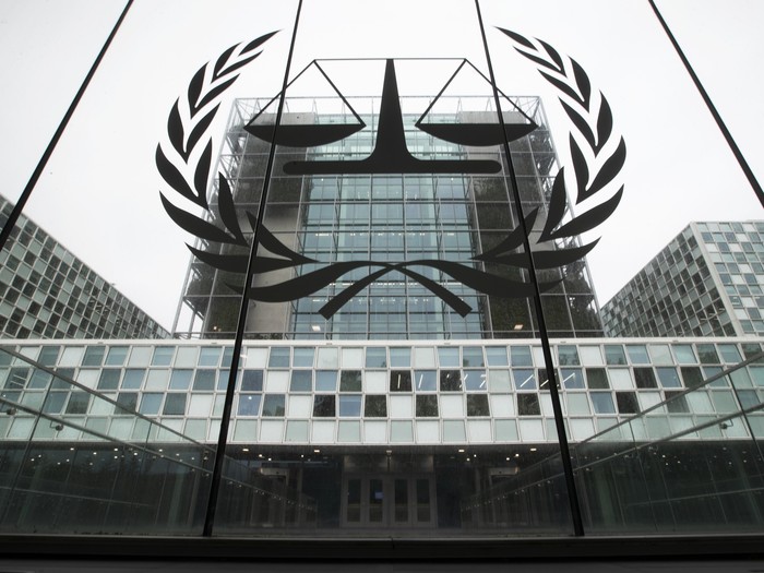 Foto durch ein Fenster auf das Gebäude des 'International Criminal Court (ICC). Auf der Fensterfront ist das Logo des Internationalen Strafgerichtshof abgebildet. Eine Waage umkreist von einem oben offenen Lorbeerkranz