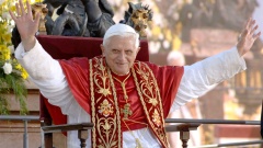 Papst Benedikt XVI. sitzt auf einem Thron und hebt die Hände zum Gruß