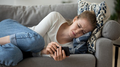 Eine Jugendliche liegt auf einem Sofa mit Handy in der Hand und ist traurig.
