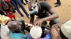 Eine Person untersucht im Südsudan auf einem freien Platz die Augen einer Person und andere schauen zu.