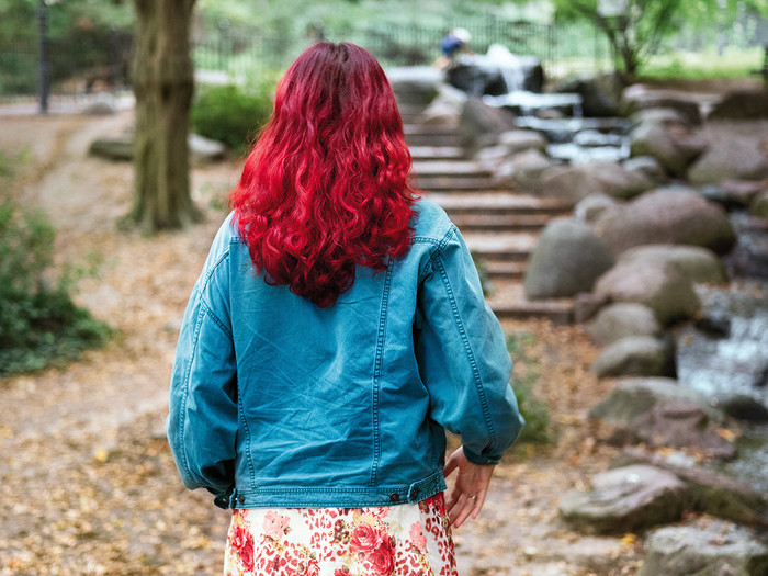 Junge Frau mit langen, rot gefärbten Haaren, läuft in einer Parkanlage an einem Bach entlang 