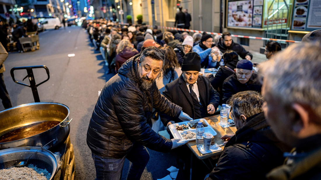 Ein Mann verteilt Essen an viele Menschen die an Tischen auf der Strasse sitzen
