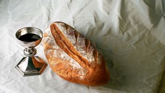 Auf einer weißen Tischdecke steht ein silberner Kelch und liegt ein Brot.