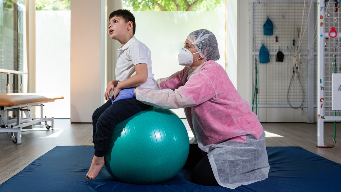 Physiotherapeutin arbeitet mit behindertem Kind im Gymnastikraum unter Corona Hygienevorschriften