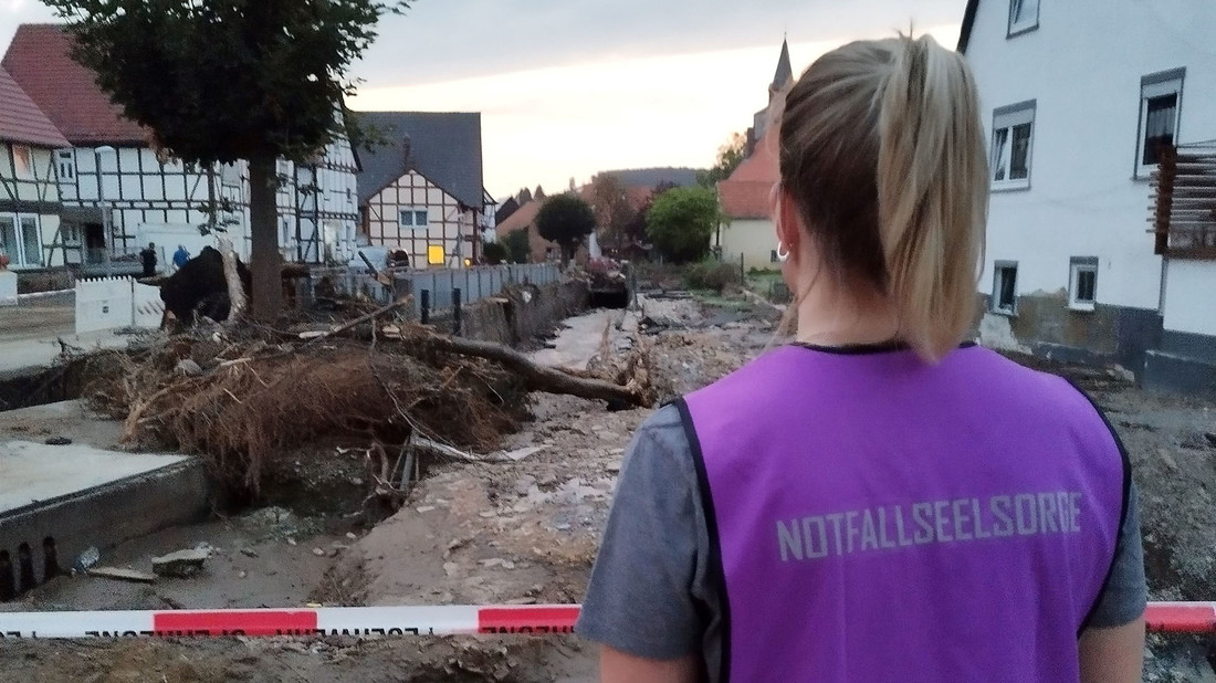 Eine Frau mit einer Notfallseelsorgeweste an schaut auf die Folgen einer Überschwemmung in einer Stadt.