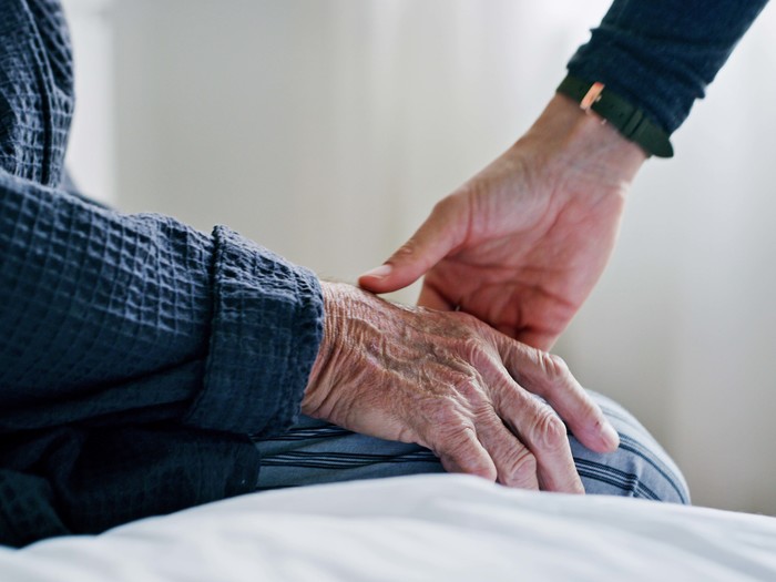 Älterer Mann sitzt auf einem Krankenbett. Seine Hand ruht auf den Beinen. Die Hand einer jüngeren Person greift nach seiner Hand