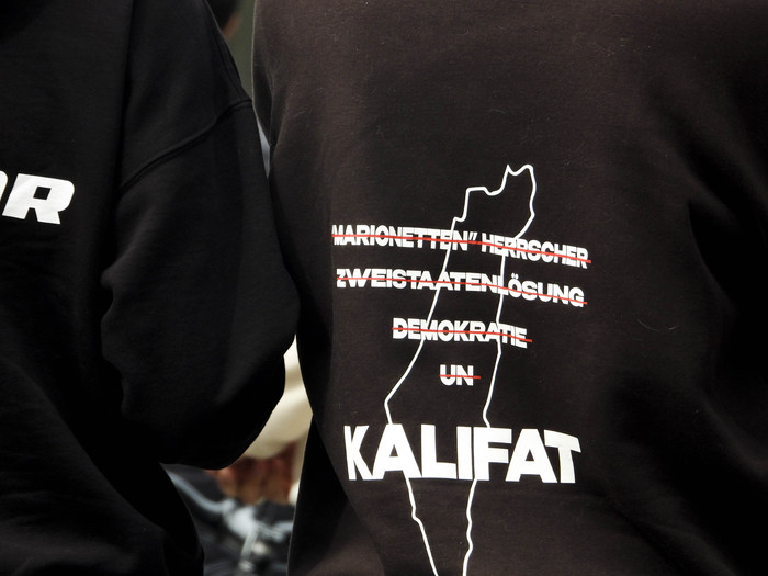 Zwei Männer tragen Sweatshirts mit der Forderung nach dem Kalifat. Die Begriffe "Marionetten Herscher, Zweistaatenlösung, Demokratie, UN" sind durchgestrichen