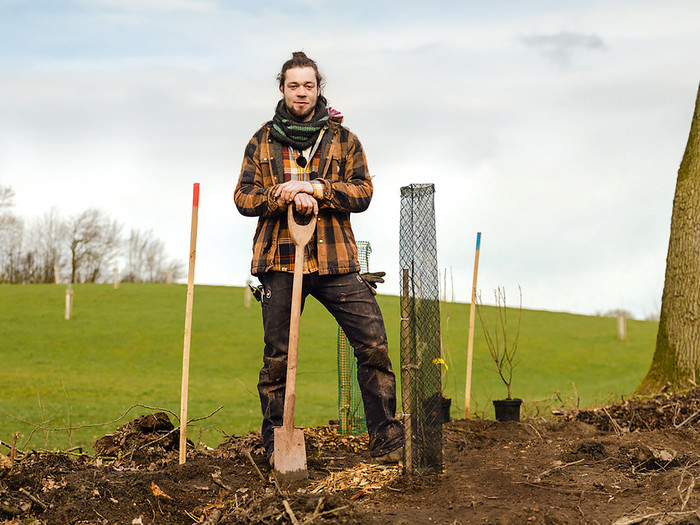 Felix Riecken steht mit einer Schaufel auf einem Stück Land und vor neu eingepflanzten Bäumen