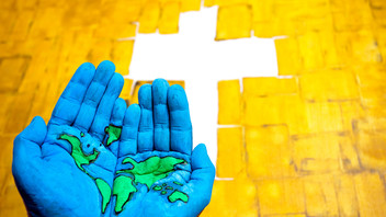 bemalte Hände mit einer Weltkarte vor einem Kreuz auf gelbem Hintergrund