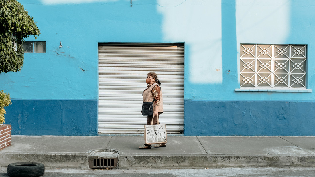 Eine Frau, die Mutter des Fotografen, läuft mit Einkaufstasche und Mundschutz vor einer blauen Wand