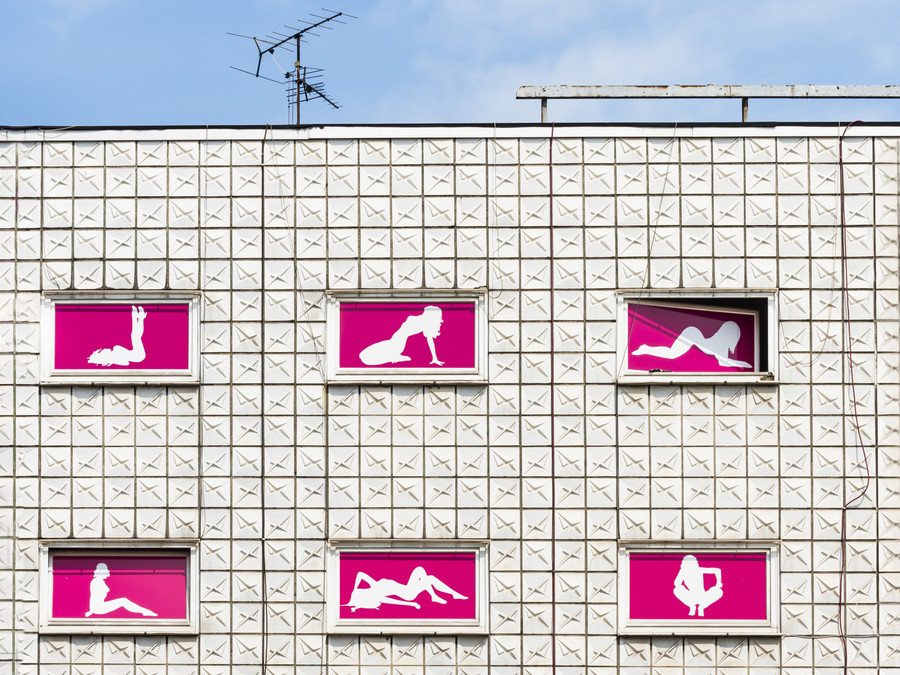 Blick auf ein Haus, dessen Fenster mit pinkfarbenen Plakaten bedeckt sind. Auf den Plakaten sind die Körper von Frauen zu sehen
