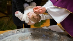 Bild von einer katholischen Taufe. Kind wird über die Taufschale gehalten, der Priester benetzt den Kopf mit Wasser.