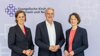 Zwei Frauen und ein Mann posieren für ein Foto vor einer Wand mit einem Schrifftzug drauf Evangelische Kirche Hessen und Nassau.