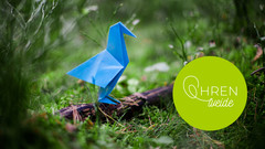 Ein blauer Papiervogel sitzt im Wald auf einem Stück Holz auf der Erde.