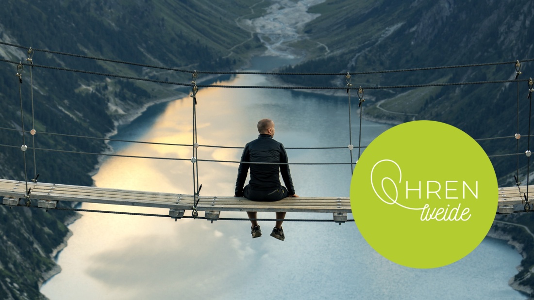 Mann sitzt auf einer Hängebrücke hoch über einem Bergsee