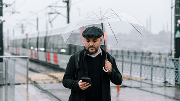 Ein Mann steht mit Regenschirm auf der Straße und hat ein Handy in der Hand.