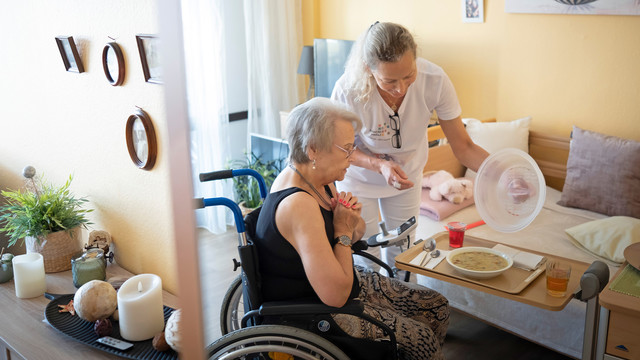 Eine Pflegerin reicht einer Seniorin im Rollstuhl das Essen