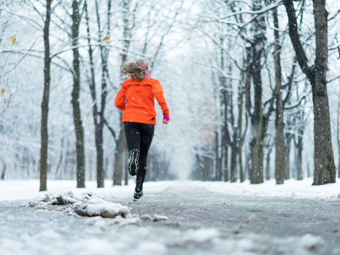 Frau mit roter Jacke joggt Frau Joggen im Winter bei Schneefall auf einer schneebedeckten Allee 