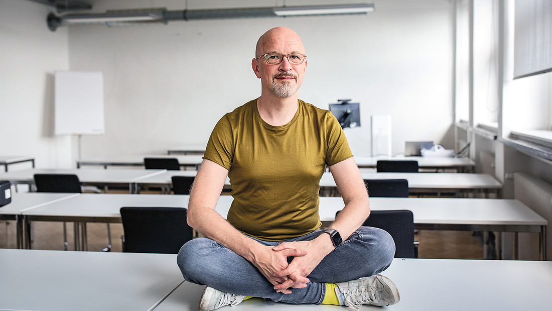 Ein Mann mit gelbem Shirt sitzt im Schneidersitz auf einem Tisch in einem Seminarraum