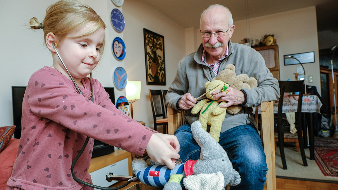 Ein Kind untersucht mit einem Stethoskop ein Stofftier und der Großvater im Hintergrund hat Teddys im Arm
