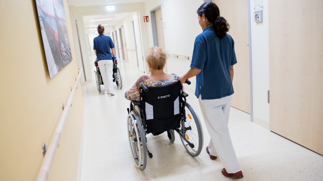 Zwei Pflegerinnen schieben Patienten mit Rollstühlen durch einen Krankenhausflur.