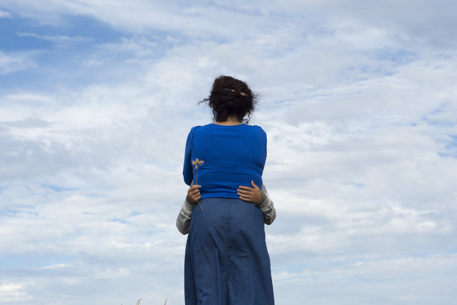 Eine Frau steht vor blauem Himmel mit dem Rücken zum Betrachter. Sie wird von einem Kind umarmt, von dem nur die Hände zu sehen sind 