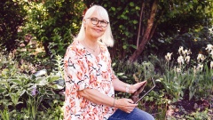 Eine blonde Frau mit Brille sitzt im Garten und hält ein Tablet in der Hand
