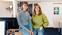 Zwei junge Frauen lehnen an einen Schreibtisch und blicken in die Kamera