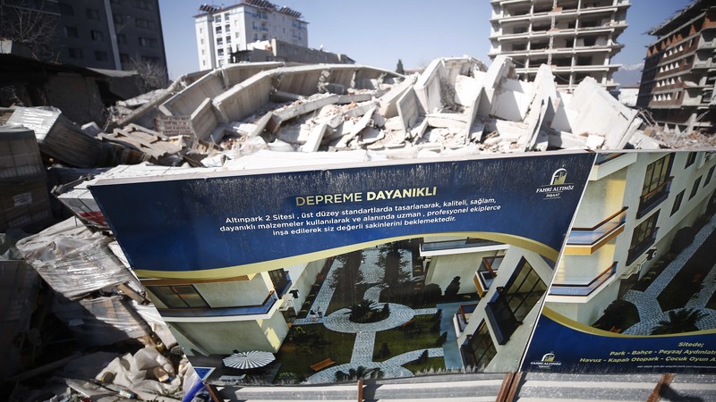 Trümmerhaufen in der Stadt Hatay. Auf einem Bauschild vor dem eingestürzten Gebäude ist zu lesen "Erdbebensichere Gebäude". 