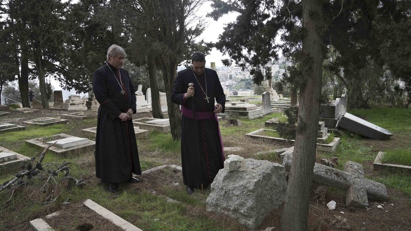 Zwei Bischöfe der Anglikanischen Kirche stehen auf dem geschändeten lutherisch-anglikanischen Friedhof auf dem Berg Zion in Jerusalem vor einem Grab mit abgebrochenem Kreuz. 