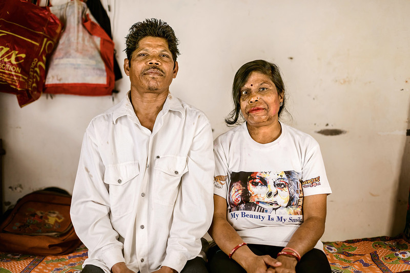 Foto von Anil (45) und Mahdu (39) in Indien. Sie sitzen vor einer verstaubten weißen Wand auf einer einfachen Pritsche 