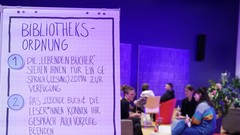 Im Vordergrund ein Schild mit Bibliotheksregeln, im Hintergrunf fünf Personen