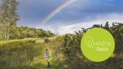 Mädchen läuft durchs grüne Feld, über ihr ein Regenbogen am Himmel