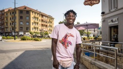 Ein lächelnder Schwarzer Mann steht in Italien auf der Straße. Er trägt ein rosa Shirt, eine rosa Gürteltasche und eine Jeans