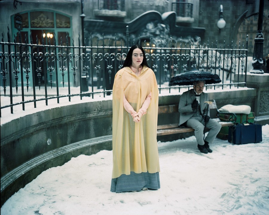 Emma Stone als Bella Baxter in "Poor Things": Frau in gelbem Umhang steht im Schnee, im Hintergrund ein Mann im Anzug mit Schirm auf einer Bank