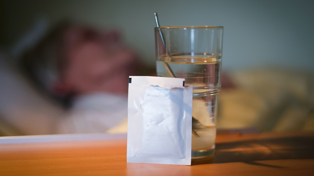 Symbolbild für Sterbehilfe mit Glas und Medikament im Tütchen neben alter Person im Bett.