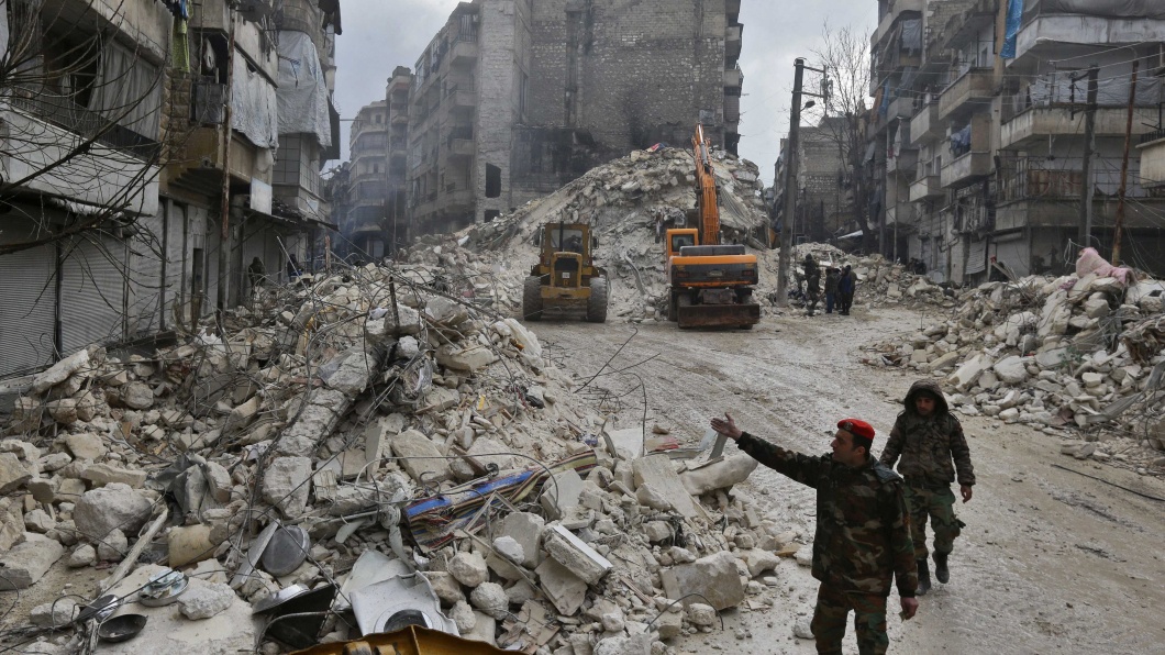 Zwei Männer von Rettungsteams laufen durch zerstörte Straßen in Aleppo, vorbei an Schuttbergen und eingestürzten Häusern.