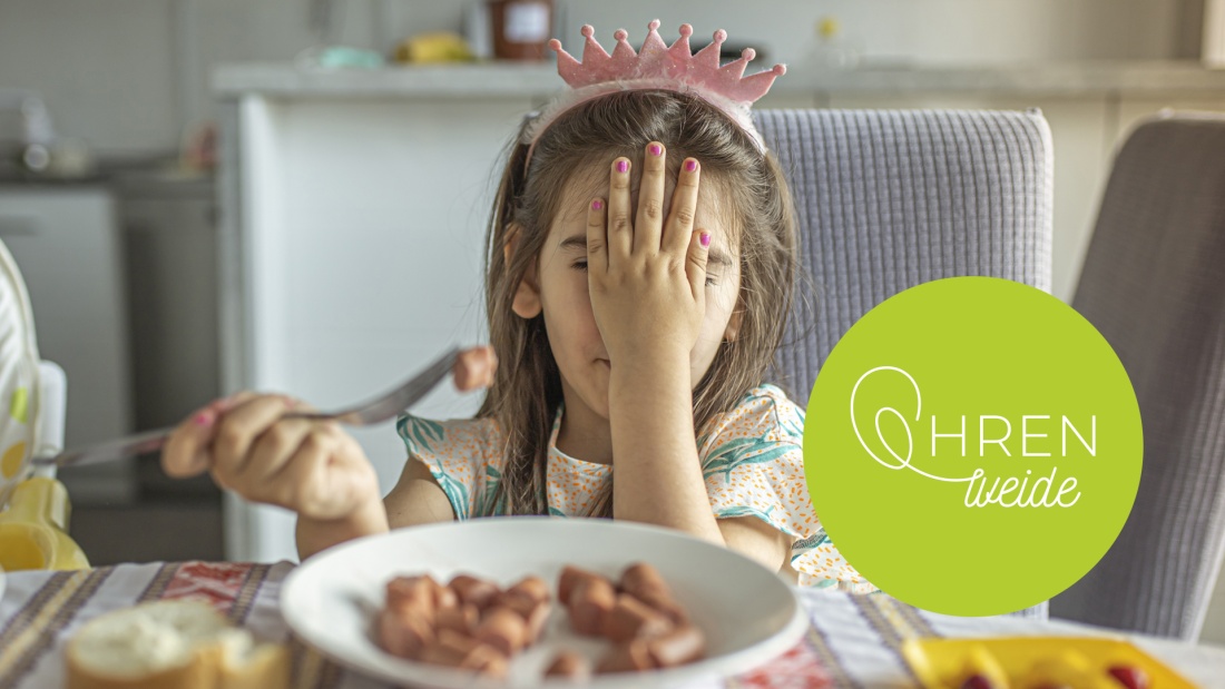 Mädchen mit pinker Krone am Essenstisch hält eine Hand vors Gesicht