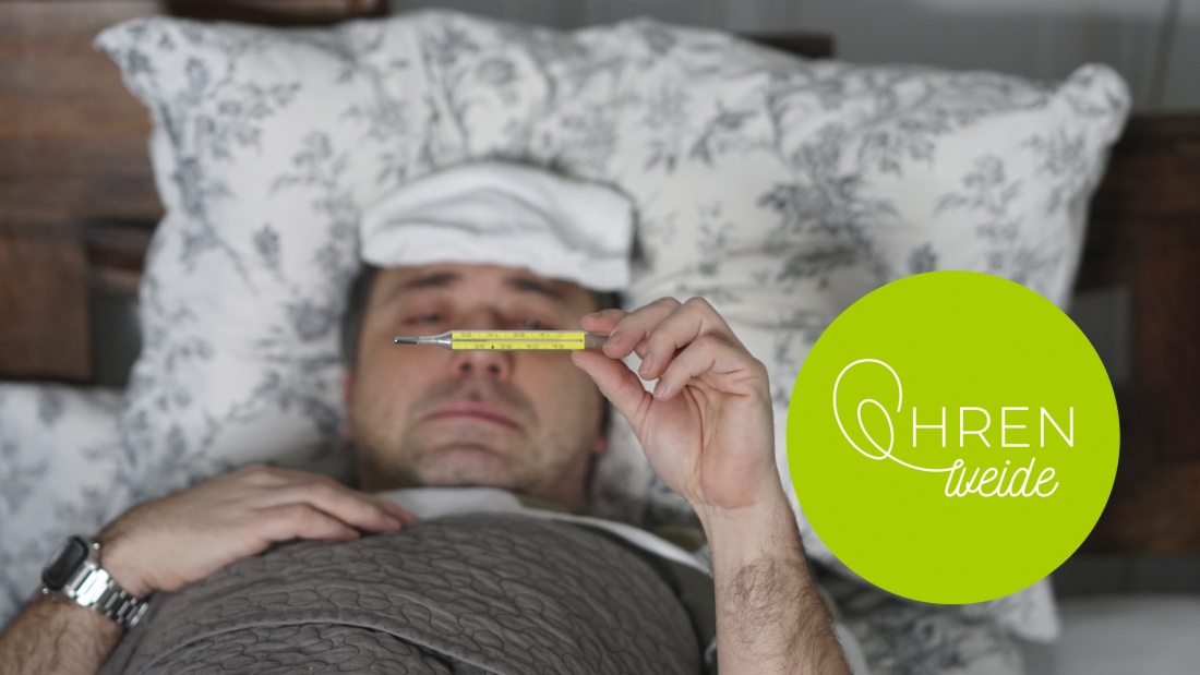 Mann liegt mit Grippe im Bett und schaut auf ein Fieberthermometer
