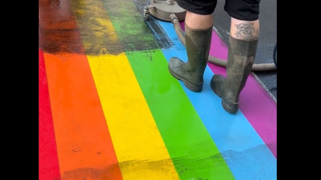 Bild zeigt, wie ein auf der Straße aufgemalter Regenbogen, den Unbekannte beschmiert hatten, gereinigt wird. Der Regenbogen war in Wiesbaden vom Dezernat für Gleichstellung anlässlich des CSD angebracht worden.